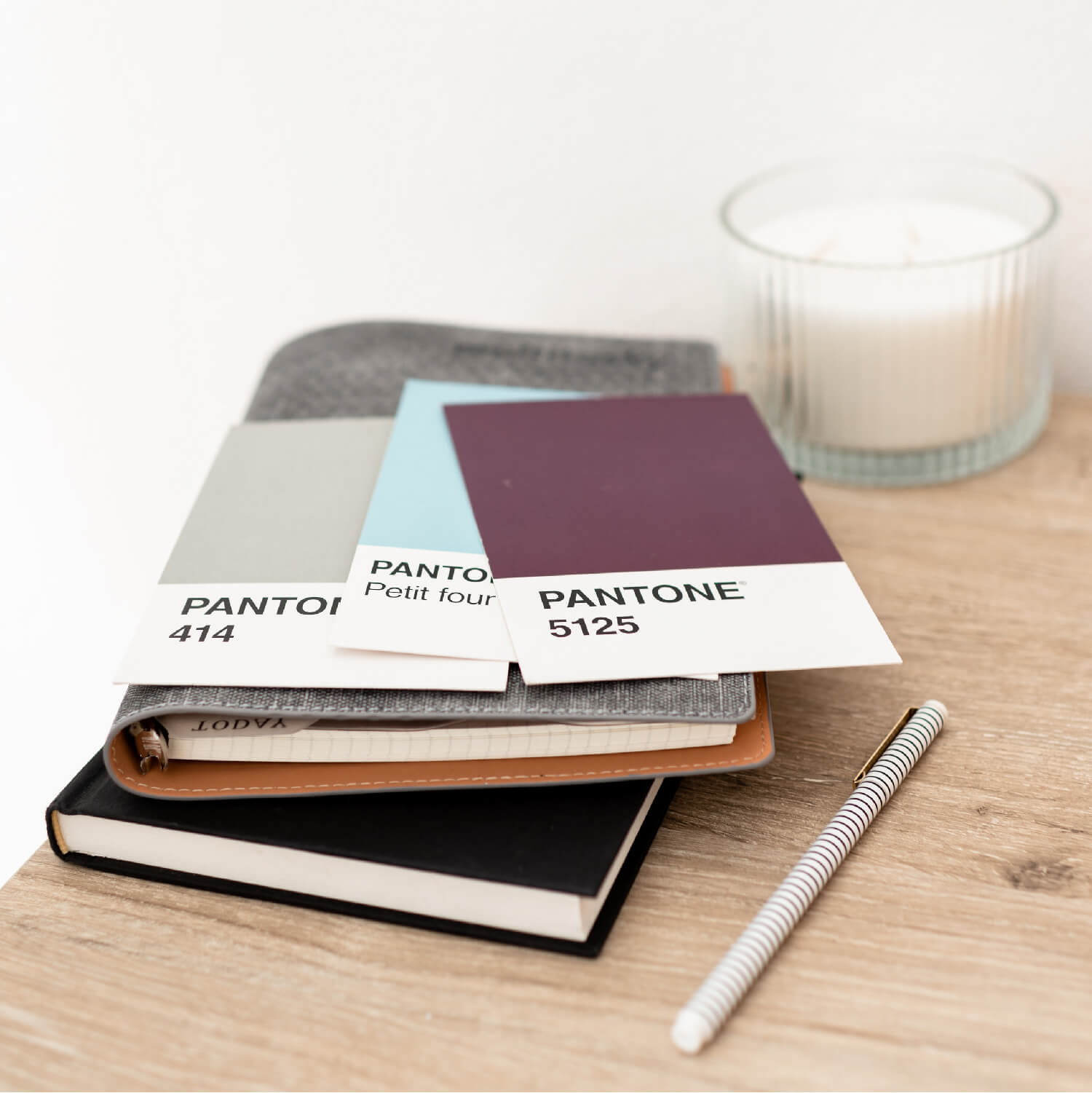 A carefully arranged workspace featuring three Pantone color swatches on top of a notebook, with a silver pen and a candle in the background, symbolizing the thoughtful process of selecting a color palette.