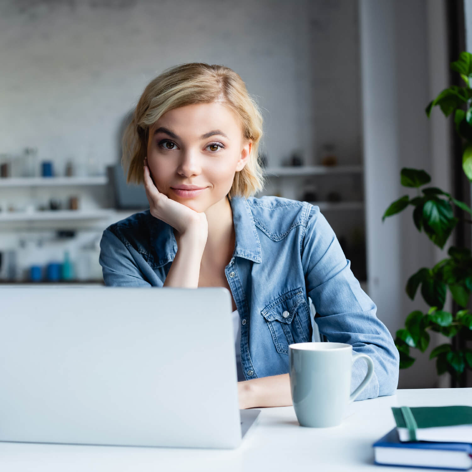 A young woman sitting at a white desk in a modern office. She is with an open laptop, a coffee mug, and notebooks in front of her, creating a friendly and approachable image suitable for a blog post on SEO essentials for small businesses.
