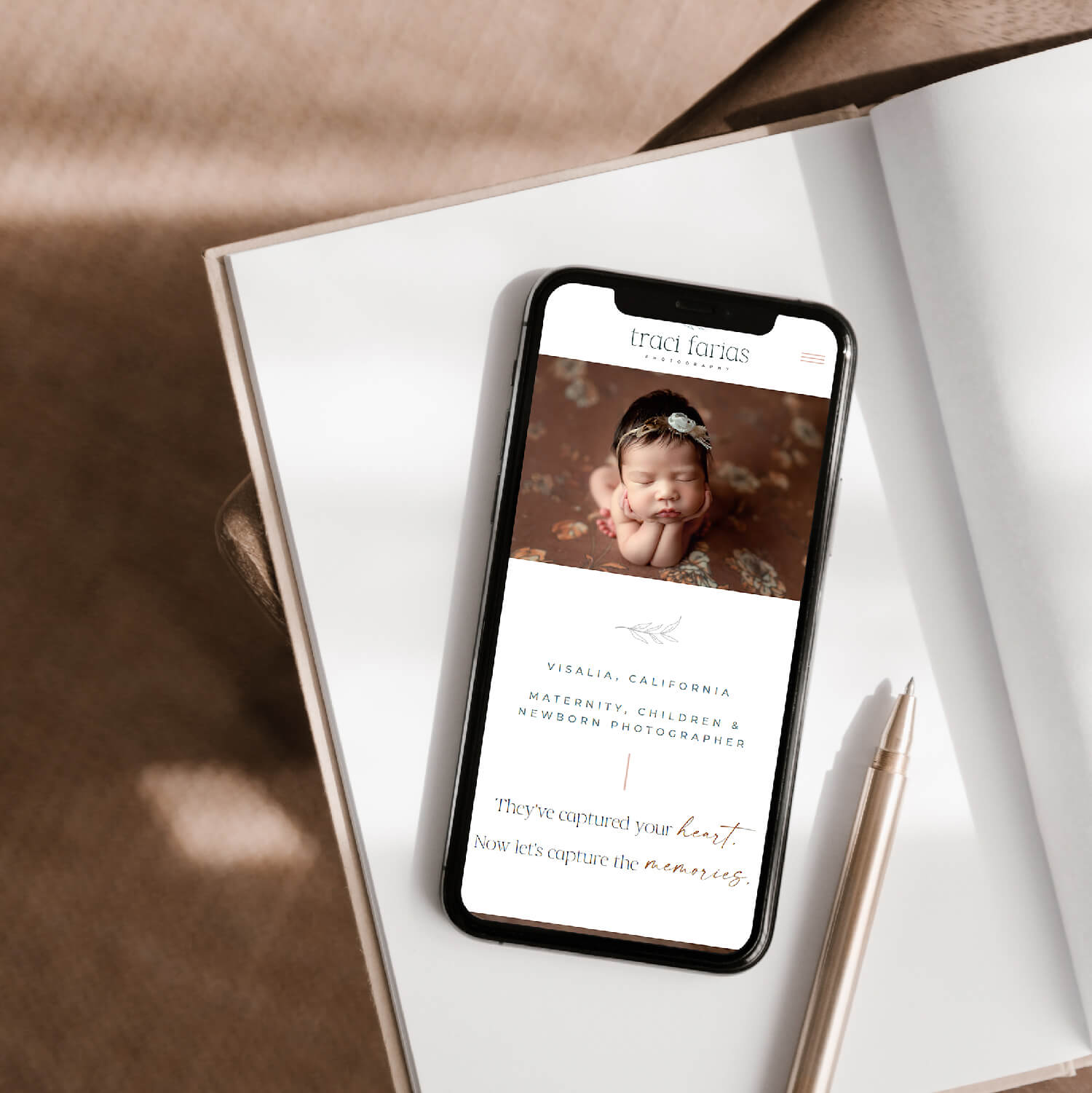 A smartphone on an open book displaying a photography website with a photo of a sleeping newborn. The site includes text for maternity, children, and newborn photography services in Visalia, California, with the tagline, 'They've captured your heart. Now let's capture the memories.'