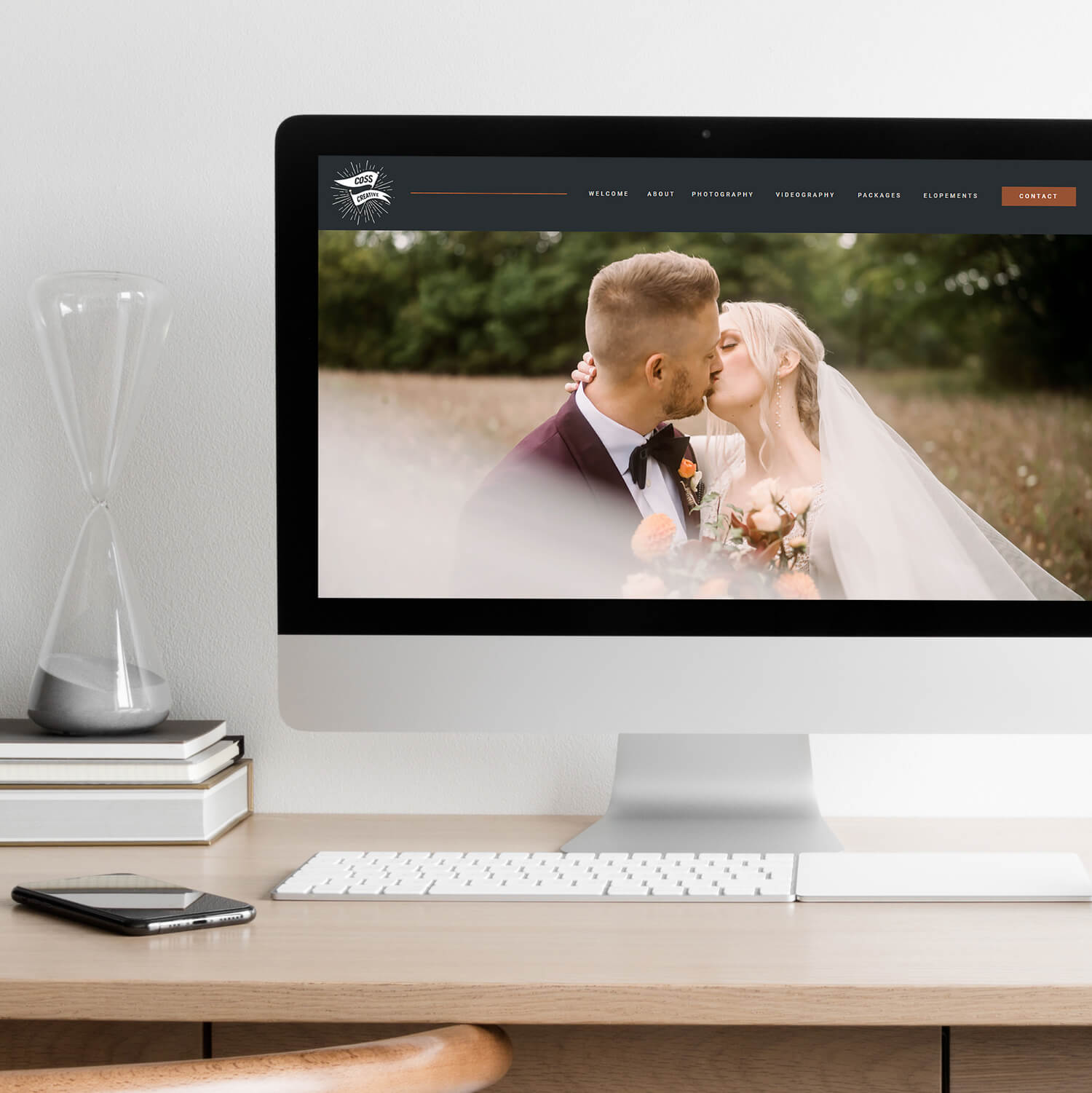 A modern home office setup with an iMac displaying a wedding photographer's website, featuring an image of a bride and groom kissing. In the foreground, a sand hourglass and a smartphone rest on the desk, enhancing the contemporary, professional atmosphere.